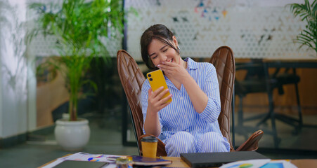 Afghan young adult gen z woman lady sitting on chair use mobile look cam talk gossip indoor job place happy girl staff hold phone speak on video call after work enjoy break spend day time at office