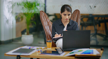 Indian gen z busy woman lady sitting chair use laptop on desk table look screen check email job...