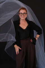 Portrait of a smiling woman. The woman is wearing a black blouse, holding glasses. The woman has red hair. The photo was taken in the studio. Fifty.