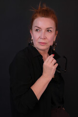 Portrait of a smiling woman. The woman is wearing a black blouse, holding glasses. The woman has red hair. The photo was taken in the studio. Fifty.