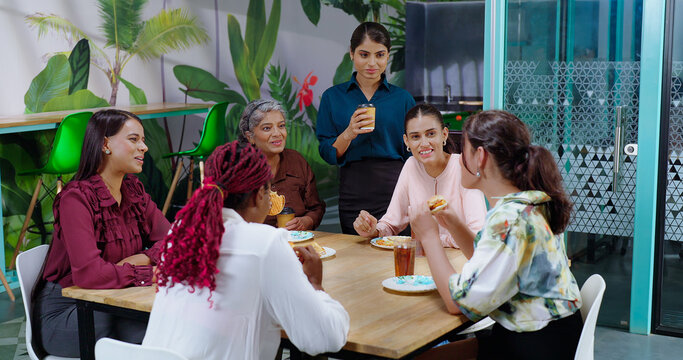 Happy asian woman girl old lady senior group team talk gossip eat sandwich share lunch indoor office work place Indian diverse female sitting around table enjoy breakfast tasty juice cup have fun joy
