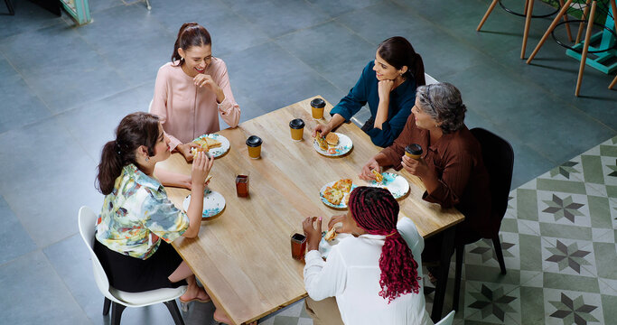 Indian happy woman staff sitting around table with old aged lady talk gossip eat tasty pizza junk food enjoy break time female girl group team speak funny joke drink coffee have fun joy at work place