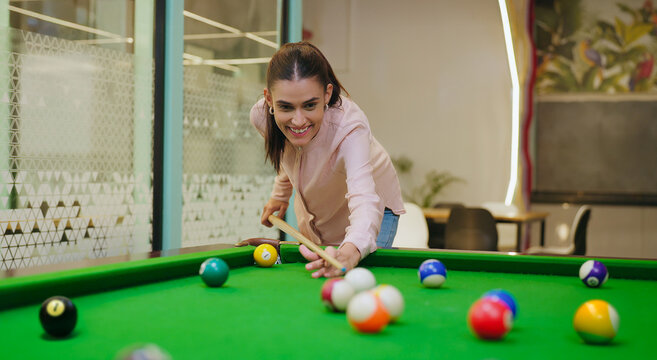Indian young adult gen z happy woman lady hold cue stick play snooker game on table pool after work at indoor office. Asian girl staff intern enjoy casual break spend good time have fun joy job place