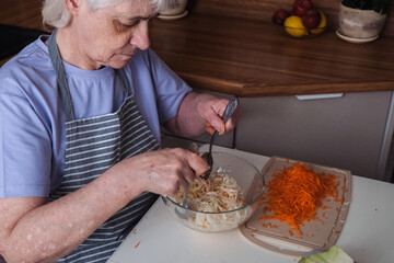 An elderly woman with short hair sit at a kitchen counter, mixing a fresh cabbage and carrot salad .