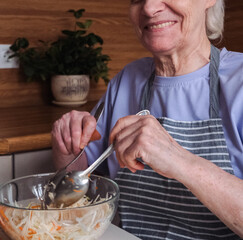 An elderly woman with short hair sit at a kitchen counter, mixing a fresh cabbage and carrot salad .
