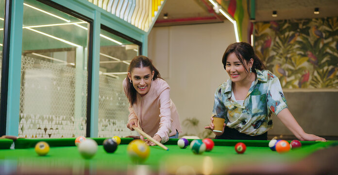Indian happy young adult gen z woman lady hold cue stick hit ball play snooker game on table office place girl staff talk drink coffee spend time have fun joy