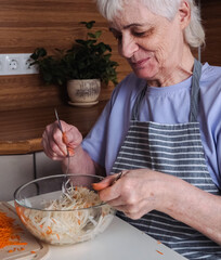 An elderly woman with short hair sit at a kitchen counter, mixing a fresh cabbage and carrot salad .