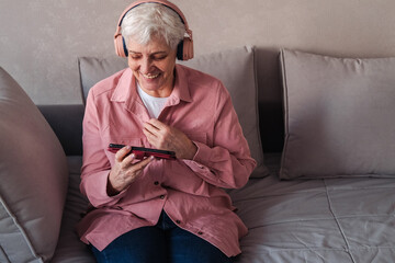 Smiling relaxed senior woman wearing headphones sitting on sofa at home using smart phone looking...