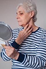 Beautiful elderly woman holding mirror and applying face cream at home