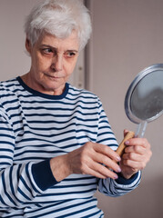 Beautiful elderly woman holding mirror and applying face cream at home