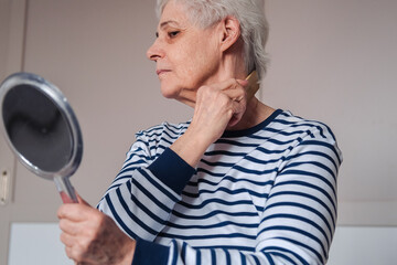 Woman Looking At Herself In The Mirror. Beautiful elderly woman holding mirror and applying face cream at home.