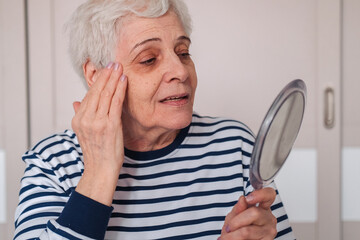 Woman Looking At Herself In The Mirror. Beautiful elderly woman holding mirror and applying face...