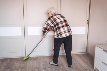 An elderly woman wearing casual clothes is sweeping the room.