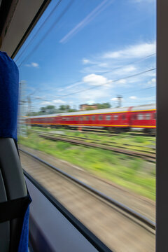 View from train window of red passenger train passing on parallel tracks with motion blur effect under blue cloudy sky, showing railway transportation system