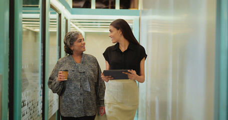 Indian older woman lady talk gossip young adult staff discuss work day time task indoor office place smiling female hold paper cup drink coffee expert girl hold tablet device walk at hall way space