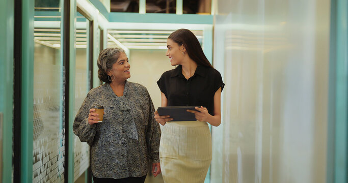 Indian older woman lady talk gossip young adult staff discuss work day time task indoor office place smiling female hold paper cup drink coffee expert girl hold tablet device walk at hall way space