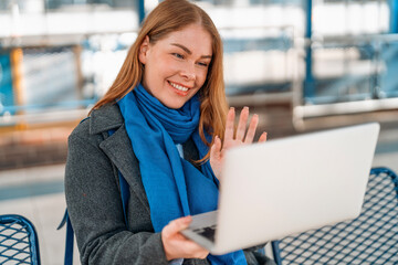 Smiling woman waves while using laptop in a public space during daytime