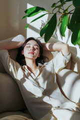 Young woman relaxing on sofa with eyes closed, enjoying sunlight and houseplant shadow