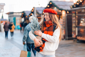 Young woman in a beret and scarf joyfully holding a small Christmas tree at a festive market during...