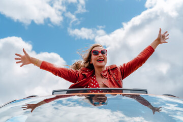 Joyful woman enjoying a road trip with arms raised in the air under a blue sky with fluffy clouds...