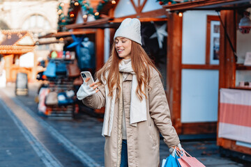 Young woman enjoys shopping at a festive market while using her phone in a charming winter setting...