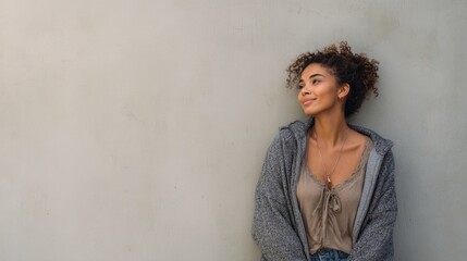 A young woman with curly hair smiles thoughtfully while leaning against a plain beige wall, dressed casually in a gray cardigan and beige top.