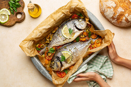 Hands holding cooking tray with baked Dorado fish or sea bream with tomatoes and olives, fresh bread, olive oil, lemon. Mediterranean food. Healthy dinner, top view.