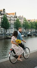 Couple Biking by Canal. A woman holding sunflowers while sitting on a bicycle, accompanied by a man, riding by a scenic canal with classic European architecture in the background.