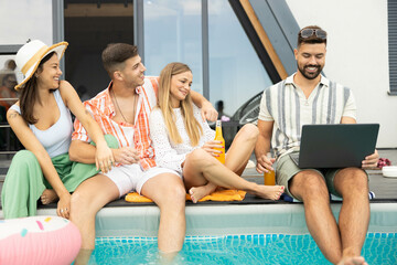 A group of four friends relax by the pool, enjoying drinks and company while one checks his laptop, blending fun with technology.