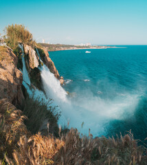  Panoramic view of Duden Waterfalls flowing powerfully into the sea from Antalya’s cliffs, with the coastal cityscape, yacht on the water, and golden grasses under clear blue sky.