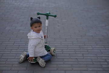 Smiling toddler boy standing outdoors with a green scooter on a paved street, wearing a striped...
