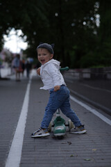 Obraz premium Smiling toddler boy standing outdoors with a green scooter on a paved street, wearing a striped hoodie, jeans, and a cute hat with ears. Urban background with soft natural lighting.