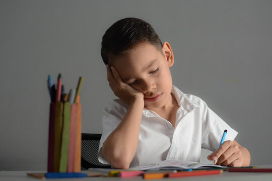 Young boy looking sad or bored while working on school homework, resting his head on his hand at a desk.
