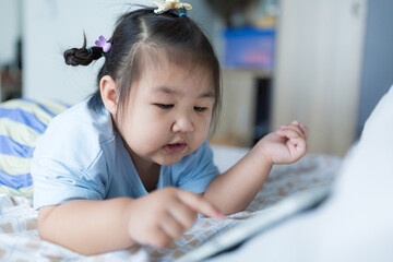 A little Asian girl is lying on the bed watching a tablet,Asian girl lying on bed in bedroom and looking at tablet screen alone with happy moment. Concept of internet, social media.
