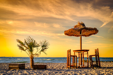 Relaxing beach setup with thatched umbrella and wooden chairs under a clear blue sky during late...