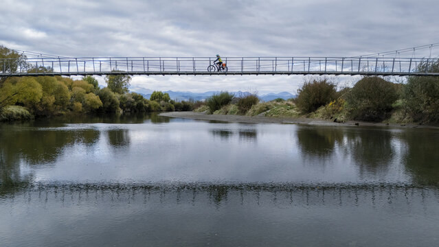 Aerial view of a cyclist crossing the CB Kidson suspension bridge over the water, reflecting the cloudy sky above the tree line, Waimea River, Tasman Region, New Zealand.