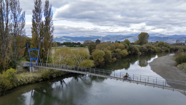 Aerial view of a cyclist crossing the CB Kidson suspension bridge over the water, reflecting the cloudy sky above the tree line, Waimea River, Tasman Region, New Zealand.