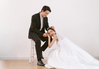 Handsome young Asian man sitting on chair and beautiful Asian bride in white dress sit on floor in...