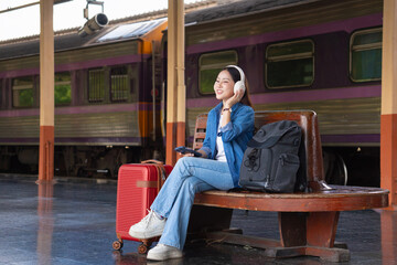 A young woman wearing headphones is listening to music on a bench in a train station.