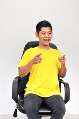 Enthusiastic Asian man in yellow shirt pointing fingers in chair with excited expression
