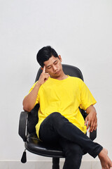 Thoughtful Asian man sitting on a chair with hand on head against white wall, lost in thought