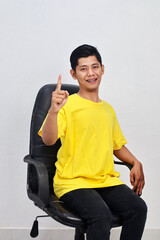 Smiling Asian Man in Yellow Shirt Sitting on Chair Pointing Upward with Positive Expression