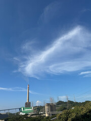 A power plant with a tall smokestack emitting smoke, set against a bright blue sky with wispy clouds.