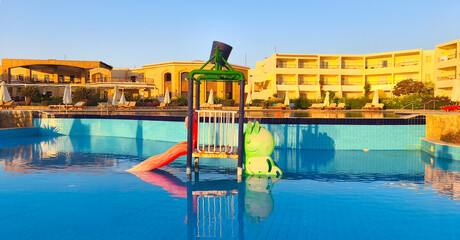 A colorful water slide and play area in a resort pool under a clear blue sky. The setting is in...