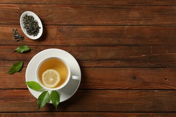 Tasty green tea with lemon in cup, brew and leaves on wooden table, flat lay. Space for text