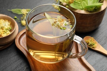 Tasty linden tea in cup, leaves and flowers on dark textured table, closeup