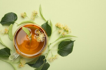 Tasty linden tea in cup, leaves and flowers on light green background, flat lay. Space for text