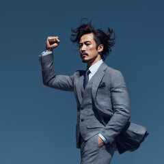 Confident Asian businessman in a stylish gray suit with long, windswept hair raising his fist in determination against a clear blue sky. The dynamic pose and professional attire convey themes of leade