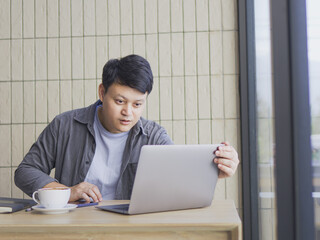 A young Asian man in casual clothes sitting at a table with a cup of coffee looking at a laptop...