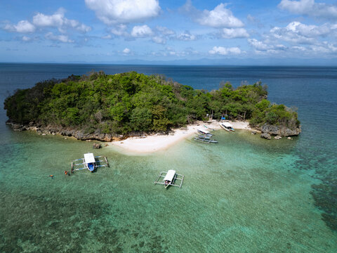 Aerial view of a lush, verdant islet embraced by the turquoise sea, with traditional boats resting on the pristine white sand, Guimaras, Western Visayas, Philippines.